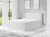 A white boxspring bed with a tufted headboard and footboard, set in a modern bedroom with large windows and artwork on the wall. Produktbild-Vorschau