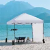 A white pop-up canopy tent set up on a sandy beach next to the ocean, with mountains in the background. A camping chair and a small cooler are placed under the tent. Produktbild-Vorschau
