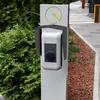 An electric vehicle charging station accessory is mounted on a post next to a green bush and a paved walkway. Náhled obrázku produktu