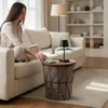 A woman sitting on a couch reaches for a phone on a side table next to a lamp. Produktbild-Vorschau