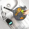 A person is cleaning a non-stick pan with multiple small circular indentations in a kitchen sink. A green sponge is being used to scrub orange residue from the pan. A black soap dispenser with a blue sponge inside is attached to the side of the sink. Náhled obrázku produktu