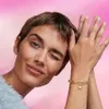 A woman winks and poses with her hands near her face, showing off a gold bracelet with a pearl charm. Produktbild-Vorschau