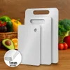 Three white cutting boards of different sizes are displayed in front of a basket of fresh vegetables and fruits, with a close-up inset showing the thickness of one of the boards. Produktbild-Vorschau