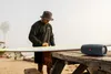 A man in a bucket hat and sweatshirt is standing on a beach next to a surfboard, with a portable speaker on a wooden table. Produktbild-Vorschau