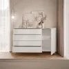 A white dresser with an open door and drawers, decorated with framed art and a vase of flowers on top, in a room with light-colored walls and curtains. Produktbild-Vorschau