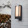 A modern LED outdoor wall light with a motion sensor is mounted on a white brick wall. Anteprima dell’immagine prodotto
