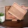 A hand is shown holding a wooden cutting board over another cutting board on a wooden table, with a potted plant in the background. Produktbild-Vorschau