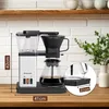 A coffee maker is shown on a wooden countertop, with a brick wall and shelves in the background. Produktbild-Vorschau