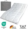 A white duvet is laid out diagonally on a white background, with a grey storage bag to the left. Several certification and information icons are present in the bottom left and right corners. Produktbild-Vorschau