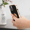 A hand holding a black phone case with a gold ring holder, next to a vase with flowers. Produktbild-Vorschau
