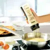 A hand holding a GEFU Crêpes-SpätzleMix TWISTER dispenser, pouring batter into a frying pan on a stovetop. Ingredients like eggs and flour are visible in the background. Produktbild-Vorschau