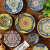 A collection of colorful, patterned plates arranged on a wooden table, with a sprig of greenery in the background. Náhled obrázku produktu