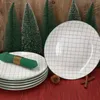 A stack of plates with a green napkin and a pinecone on a red tablecloth, with decorative Christmas trees in the background. Náhled obrázku produktu