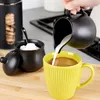 A hand is pouring milk from a black creamer into a yellow mug of coffee. A black sugar bowl with a spoon and a black cookie jar are also visible. Produktbild-Vorschau