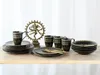 A set of black and gold patterned tableware, including mugs, plates, and bowls, arranged on a wooden surface with a decorative statue in the background. Náhľad obrázku produktu