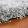 A close-up view of the edge of a shaggy, high-pile rug with silver or gray fibers resting on a brown surface. Produktbild-Vorschau