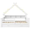 A white children's house-shaped bed with a slatted base, a headboard with shelves, a guardrail, and two drawers underneath. It is decorated with string lights along the roof. Produktbild-Vorschau