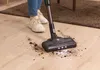 A person is vacuuming dirt off a wooden floor with a cordless vacuum cleaner. Voir les images du produit