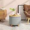 A round ottoman with a wooden top sits on a patterned rug next to a wooden cabinet and a brown armchair. A vase with a plant and a cup of cookies are on the ottoman. Produktbild-Vorschau