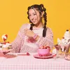 A young woman with braided hair sits at a table with a pink checkered tablecloth, surrounded by desserts and sweets, wearing a pink knitted sweater and a pink watch. Produktbild-Vorschau