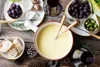 A table setting for a cheese fondue meal, with a fondue pot in the center, surrounded by bread, grapes, Brussels sprouts, and a glass of red wine. Produktbild-Vorschau