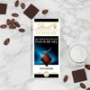 A Lindt Excellence chocolate bar with Fleur de Sel is displayed on a marble surface, surrounded by cocoa beans and a glass of milk. Produktbild-Vorschau