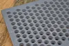 A close-up shot of a gray metal grate with many circular holes, resting on a wooden surface. Voir les images du produit