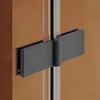 A close-up view of a black hinge on a glass shower enclosure, mounted on a clear glass panel and a brown wall. Produktbild-Vorschau