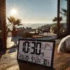 A digital weather station clock is placed on a wooden table outdoors, with a scenic view of mountains and the sun in the background. Produktbild-Vorschau
