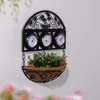 A black metal wall-mounted planter with three thermometers and a basket filled with green plants and pink flowers. Náhled obrázku produktu
