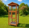 A wooden bird aviary stands on a green lawn in a garden setting with trees and a blue sky in the background. Produktbild-Vorschau