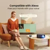 A woman is sitting on a chair holding a mug, while a projector and a smart speaker are on a coffee table in front of her. The image highlights the projector's compatibility with Alexa. Náhled obrázku produktu