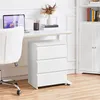 A white three-drawer nightstand is placed next to a white desk with a laptop and a vase of dried flowers. Produktbild-Vorschau