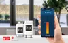 A hand holding a smartphone displaying a smart home app, with two smart thermostats in the foreground, placed on a table in front of a window. Produktbild-Vorschau