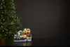 A miniature Christmas village scene with a house and trees, lit by LEDs, placed on a dark surface in front of a blurred Christmas tree. Podgląd zdjęcia produktu
