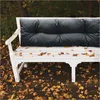 A white garden bench with a black cushion sits outdoors in autumn, surrounded by fallen leaves. Podgląd zdjęcia produktu