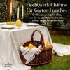 A picnic basket filled with lemons, bread, and a bottle of wine sits on the grass next to a table set for a garden lunch. Náhľad obrázku produktu
