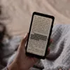 A person is holding an e-reader device and reading a book on it, with a cozy blanket in the background. Náhled obrázku produktu