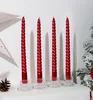 Four red spiral candles in clear holders on a white surface, with a pinecone and a hint of a Christmas tree on the right. Náhled obrázku produktu