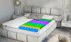 A cutaway view of a mattress topper showing its layered, multi-colored foam structure, placed on a bed in a bedroom setting. Produktbild-Vorschau