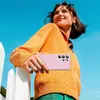 A young woman holding a pink smartphone, leaning on a white fence with a bright blue sky in the background. Produktbild-Vorschau