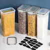 Four clear storage containers filled with pasta and grains, along with a set of black labels and a chalk marker, are displayed on a white countertop against a blue background. Produktbild-Vorschau