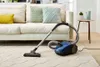 A blue Philips vacuum cleaner is placed on a cream-colored rug in a living room, with a sofa in the background. Produktbild-Vorschau