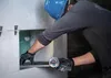 A person wearing a blue hard hat and black gloves is using a small angle grinder to cut a piece of material. The person is working inside a recessed area of a wall. Produktbild-Vorschau