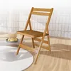 A wooden folding chair is placed on a light wooden floor, with a circular white rug in the foreground and a patterned screen or wall in the background. Produktbild-Vorschau