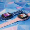 Two children's smartwatches, one blue and one pink, are displayed on a textured blue and pink surface. Náhled obrázku produktu