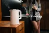 A woman in a blazer holds a drink while leaning against a kitchen counter, with a white electric kettle steaming on a wooden cabinet in the foreground. Produktbild-Vorschau