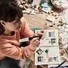 A woman is sitting on the floor, surrounded by craft supplies, and is using a glue gun to attach a photo to a scrapbook. Produktbild-Vorschau