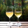 Two glasses of white wine are placed on a wooden table outdoors, with a blurred green and brown background of trees. Produktbild-Vorschau