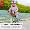 A young girl is riding a green balance bike with a basket of flowers on the handlebars. She is wearing a pink sweater and a white and pink helmet. The background is a blurred outdoor setting with greenery and a stone wall. Náhled obrázku produktu